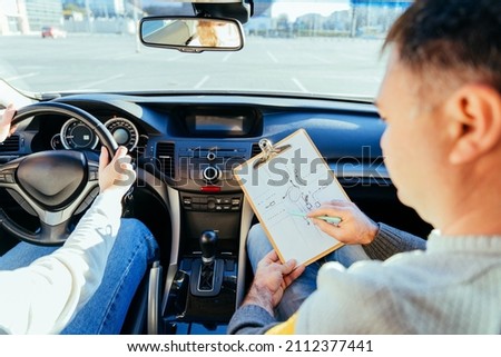 Man instructor teaching student about traffic rules on first automobile driving class. Examiner sitting with her student inside a car. Writing points. Driving instructor writing points Royalty-Free Stock Photo #2112377441