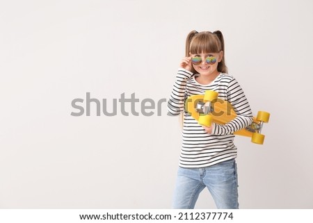 Cute little girl with stylish sunglasses and skateboard on light background