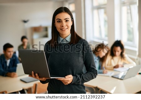 Smiling teacher using laptop during computer class at high school and looking at camera. Her students are learning in the background.  Royalty-Free Stock Photo #2112392237