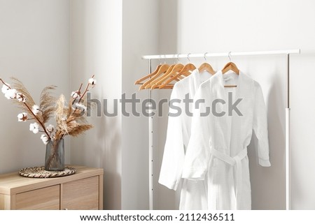 Rack with white bathrobes near light wall in room Royalty-Free Stock Photo #2112436511