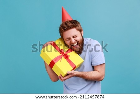 Portrait of happy positive handsome bearded man in party cone embracing yellow gift box, getting present from friend on his birthday. Indoor studio shot isolated on blue background.
