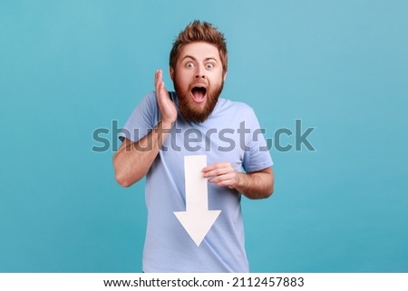 Portrait of astonished surpised bearded man showing white arrow pointing down, expressing shocked emotions, downgrade concept. Indoor studio shot isolated on blue background. Royalty-Free Stock Photo #2112457883