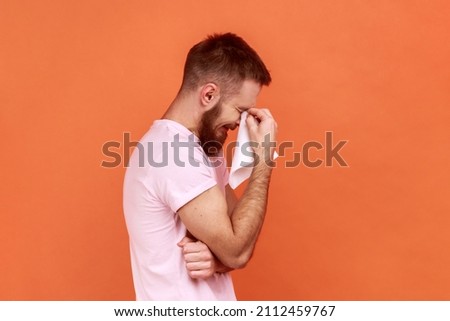 Portrait of sad alone hopeless bearded man standing, holding his head down and crying, wipes tears with handkerchief, wearing pink T-shirt. Indoor studio shot isolated on orange background. Royalty-Free Stock Photo #2112459767