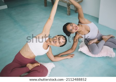 Multiracial women holding hands and practicing yoga at home. Concept of healthy lifestyle. Idea of friendship. Young smiling asian and caucasian girlfriends wear sportswear on pillows look at camera