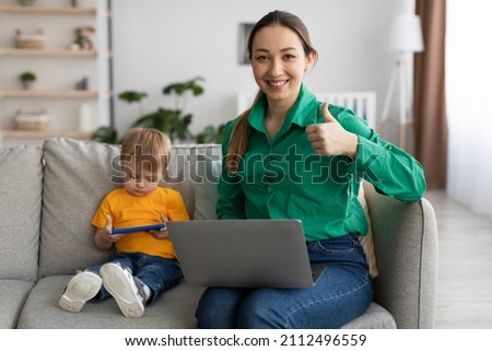 Maternity and career concept. Portrait of happy young mother working on laptop and showing thumb up, sitting near child boy watching videos on cellphone