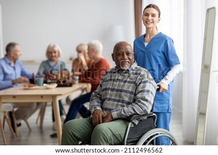 Happy black man older patient on wheelchair with female nurse smiling at camera, group of senior people sitting on couch on background, nursing home interior, healthcare for elderly people concept Royalty-Free Stock Photo #2112496562