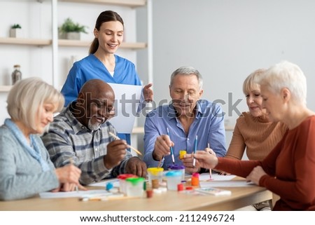 Happy multiracial elderly men and women sitting around table and drawing at nursing home, doing arts and crafts together, young lady nurse assisting group of senior people, copy space Royalty-Free Stock Photo #2112496724