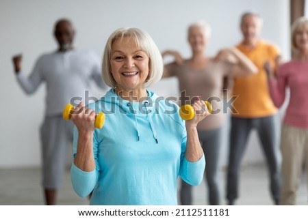Multiracial group of senior people in sportswear doing strength building fitness exercises with dumbbells, holding fitness tools and smiling at camera, selective focus on positive elderly lady Royalty-Free Stock Photo #2112511181