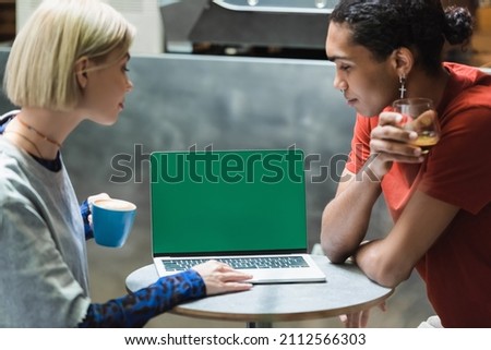 Side view of multiethnic freelancers holding coffee and using laptop with green screen in cafe