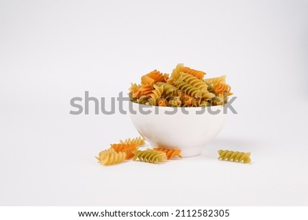 Short, extruded, twisted colored pasta on white background with copy space. Three Colors Rotini or Fusilli Pasta in white bowl and scattered on surface Royalty-Free Stock Photo #2112582305