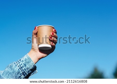 Woman hands holding a cup of coffee over blue sky background. Hipster girl holding paper cup with coffee take away. Paper coffee cup in woman hands with perfect manicure. Royalty-Free Stock Photo #2112616808