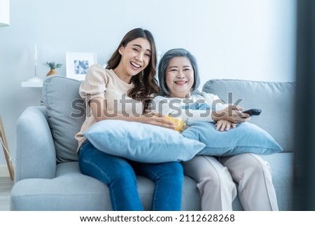 Asian senior mature woman and daughter sit on sofa watch funny movie. Loving elderly older mother spend time in living room at home with beautiful girl enjoy comedy show on TV and eat snacks together. Royalty-Free Stock Photo #2112628268