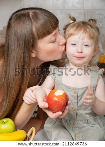 Healthy food at home. Happy family in the kitchen. Mother and little daughter cook vegetables and fruits, play. A little girl is sitting on a table in the kitchen. Mom kisses and hugs her daughter