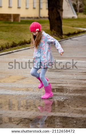 A cute little girl in a blue cape, pink boots and a pink hat runs through puddles and has a fun. Happy childhood. Early spring. Emotions.