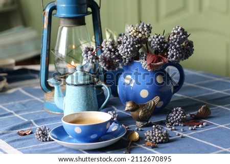 Cup of coffee, blue berries in pitcher, bronze figurine of bird, enamel coffeepot, old oil lamp on table, vintage authentic style Royalty-Free Stock Photo #2112670895