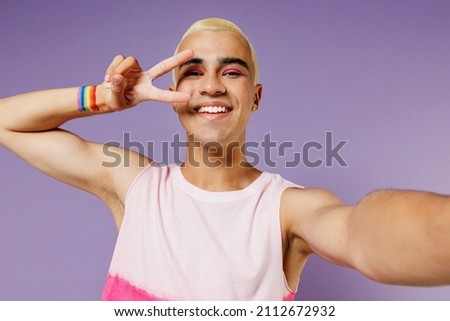 Close up young latin gay man with make up wearing bright pink top doing selfie shot pov on mobile phone show v-sign isolated on plain pastel purple background. People lifestyle fashion lgbtq concept