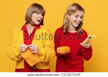 Indignant sad caucasian woman 50s in red shirt have fun with teenager girl 12-13 years old. Grandmother granddaughter knitting use mobile cell phone isolated on plain yellow background Family concept