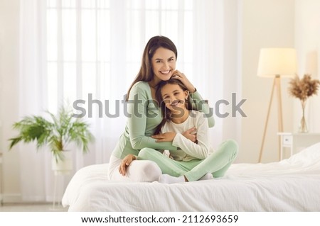 Happy family enjoying lazy morning at home. Portrait of smiling mother and child in bedroom. Beautiful mum and daughter in comfortable white and pastel green lounge homewear cuddling on cosy, warm bed Royalty-Free Stock Photo #2112693659