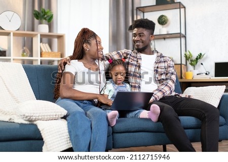 African american parents looking on each other with smile on face while cute little daughter sitting among them with laptop on knees. Family tie at home with modern technology.