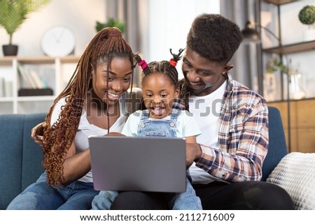 African american married couple sitting with little daughter on comfy couch and using modern laptop. Happy young family watching cartoons during leisure time at home.