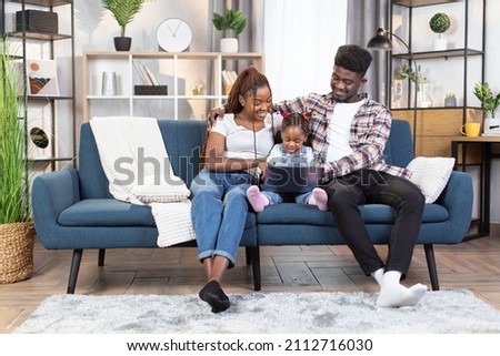 Happy african american parents with cute daughter using wireless laptop while sitting on couch. Family watching cartoons during leisure time at cozy apartment.