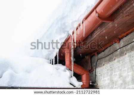A snowdrift on the roof of a house from which icicles hang. Cleaning roofs from accumulated snow. Royalty-Free Stock Photo #2112722981