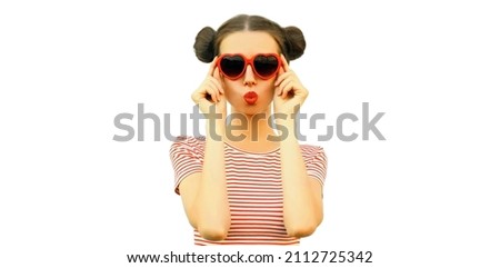 Portrait of beautiful young woman blowing her lips sending sweet air kiss with lipstick wearing red heart shaped sunglasses, striped t-shirt isolated on white background