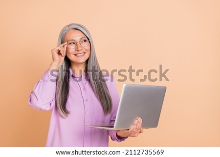 Photo of cute grey hairdo old lady hold laptop look promo wear eyewear reflecting screen purple shirt isolated on beige background