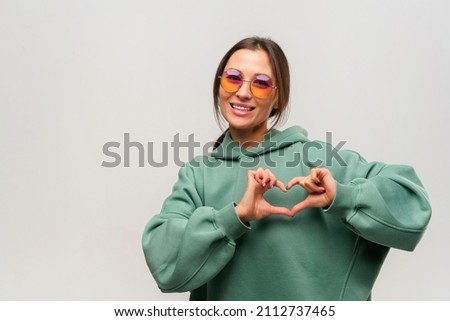 Love you. Portrait of attractive romantic young woman with brown hair standing and making heart with hands, smiling playfully. Indoor studio shot isolated on white background