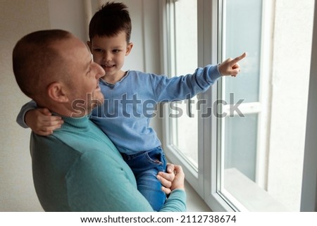 Little boy in father arms, they are standing and looking through the window at home during the day. Single Caucasian father carrying his son while looking through the window. Cute boy in father's arms