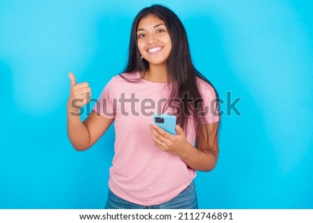 Portrait of Young beautiful woman wearing pink T-shirt against blue background using and texting with smartphone  happy with big smile doing ok sign, thumb up with fingers, excellent sign Royalty-Free Stock Photo #2112746891
