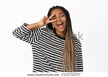 Portrait of happy and positive black girl shows peace, v-sign and winking, smiling carefree, posing against white background
