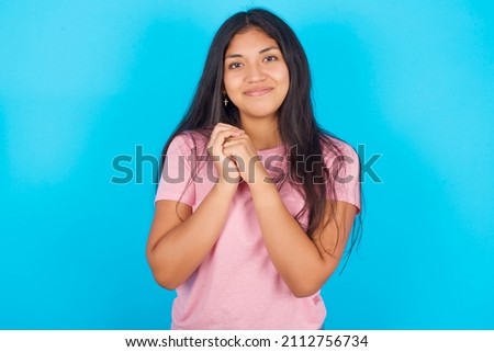Charming serious Young beautiful woman wearing pink T-shirt against blue background keeps hands near face smiles tenderly at camera