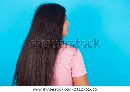 The back side view of a Young beautiful woman wearing pink T-shirt against blue background . Studio Shoot.