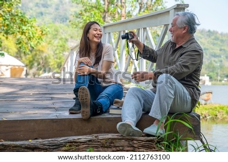 Happy Asian senior man using digital camera photography with beautiful woman in nature public park. Elderly retirement male with daughter enjoy outdoor activity together. Family health care concept