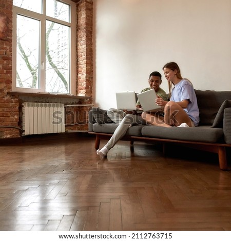 Young multiethnic couple looking on laptop of each other during working at home. Concept of freelance and remote work. Black man and caucasian woman sitting on sofa. Idea of creative generation Z