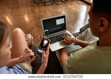Multiethnic blogger couple editing video on digital devices for social networks. Video blogging and internet multimedia. Creative girl and guy of zoomer generation creating content on sofa at home