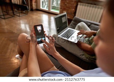 Partial image of blogger couple editing video on digital devices for social networks. Blogging and internet multimedia. Creative girl and black guy of generation Z creating content on sofa at home