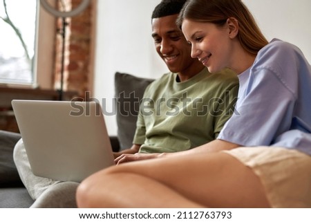 Young smiling multiracial blogger couple editing video content on laptop for social networks. Video blogging and internet multimedia. Caucasian girl and black guy of generation Z on sofa at home