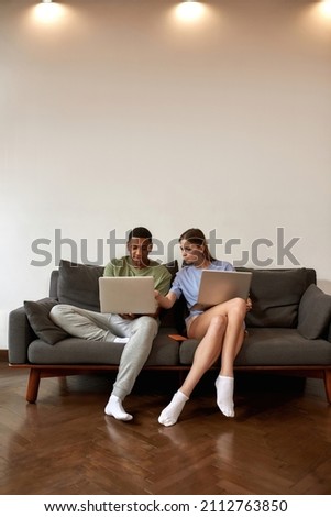 Caucasian girl pointing on laptop computer of her black boyfriend. Young multiracial couple working on sofa at home. Concept of freelance and remote work. Idea of creative zoomer generation