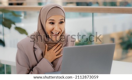 Happy young arab woman sitting at desk receiving email on laptop with good news shocked excited girl rejoices in victory making yes gesture winning achievement credit approval hired great exam result Royalty-Free Stock Photo #2112873059