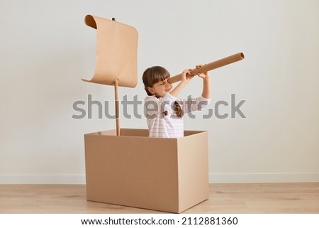 Portrait of positive funny little girl sitting in cardboard box with paper flag, playing ship captain, pretending she is in the sea, holding big spotting scope, looking far. Royalty-Free Stock Photo #2112881360