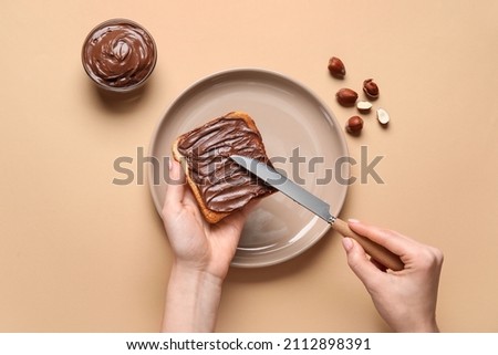 Woman preparing tasty toast with chocolate paste and hazelnuts on beige background Royalty-Free Stock Photo #2112898391