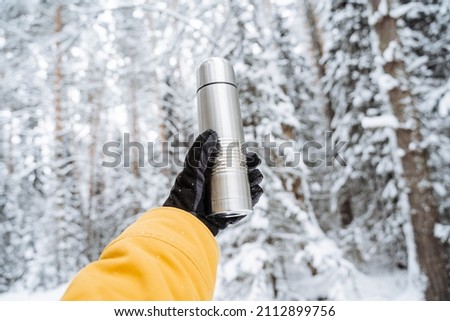 Hand with a thermos against the background of the forest in winter. Steel thermos. Metal thermo. Snow-covered white forest, taiga in the snow. Outstretched arm. High quality photo