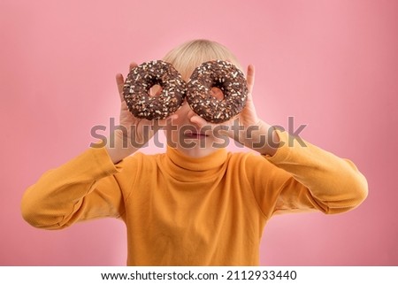 Blond child in an orange jacket holds two chocolate doughnuts. Portrait of boy with donuts on pink background