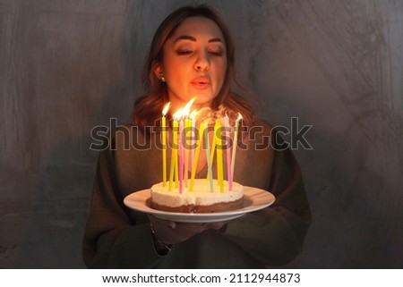 Young smiling woman holding homemade birthday cake with burning candles while standing indoors, side view. Female bringing Bday pie. Birthday traditions and celebration concept