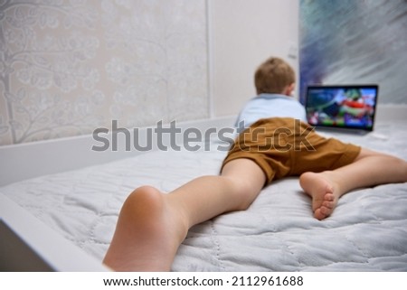 Child is watching entertainment content on a portable device while lying on the bed. A small blond boy in white wireless headphones uses a modern laptop on the bed.