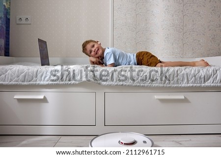 Small blond child uses a laptop and wireless headphones lying on the bed and smiling at the camera while a robot vacuum cleaner cleans his room. Modern technologies in everyday life