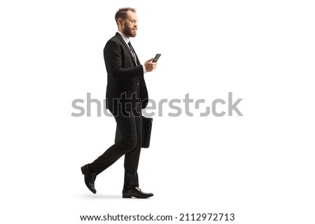Full length profile shot of a young businessman walking and using a mobile phone with a briefcase in his hands isolated on white background Royalty-Free Stock Photo #2112972713