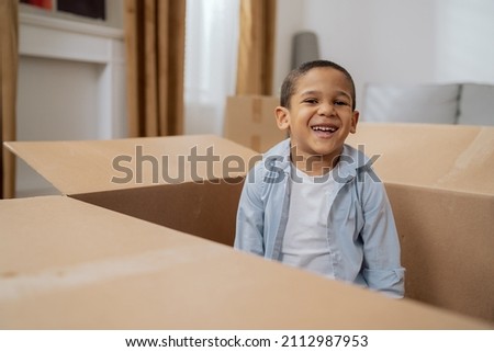Cute little boy smiling sits in cardboard box used to pack things during move, brother hides from sister for fun